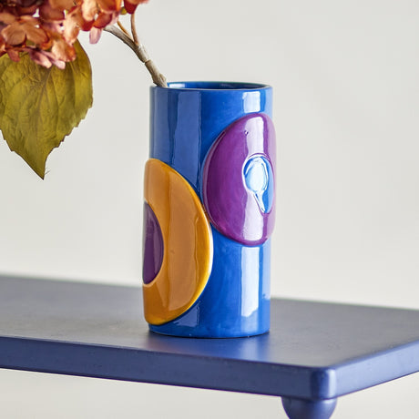 Polivia Ceramic Vase 18cm in Multi, a cylindrical high-gloss dolomite vase with raised purple and mustard geometric shapes on a blue base, shown on a dark blue shelf with dried flowers.