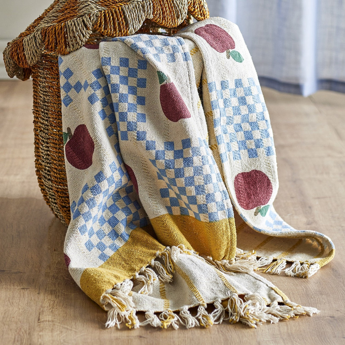 Gala Cotton Throw in Multi, a cotton throw printed with apples and blue check panels, finished with a mustard border and knotted fringe, draped over a woven basket chair.