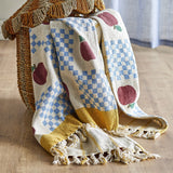 Gala Cotton Throw in Multi, a cotton throw printed with apples and blue check panels, finished with a mustard border and knotted fringe, draped over a woven basket chair.