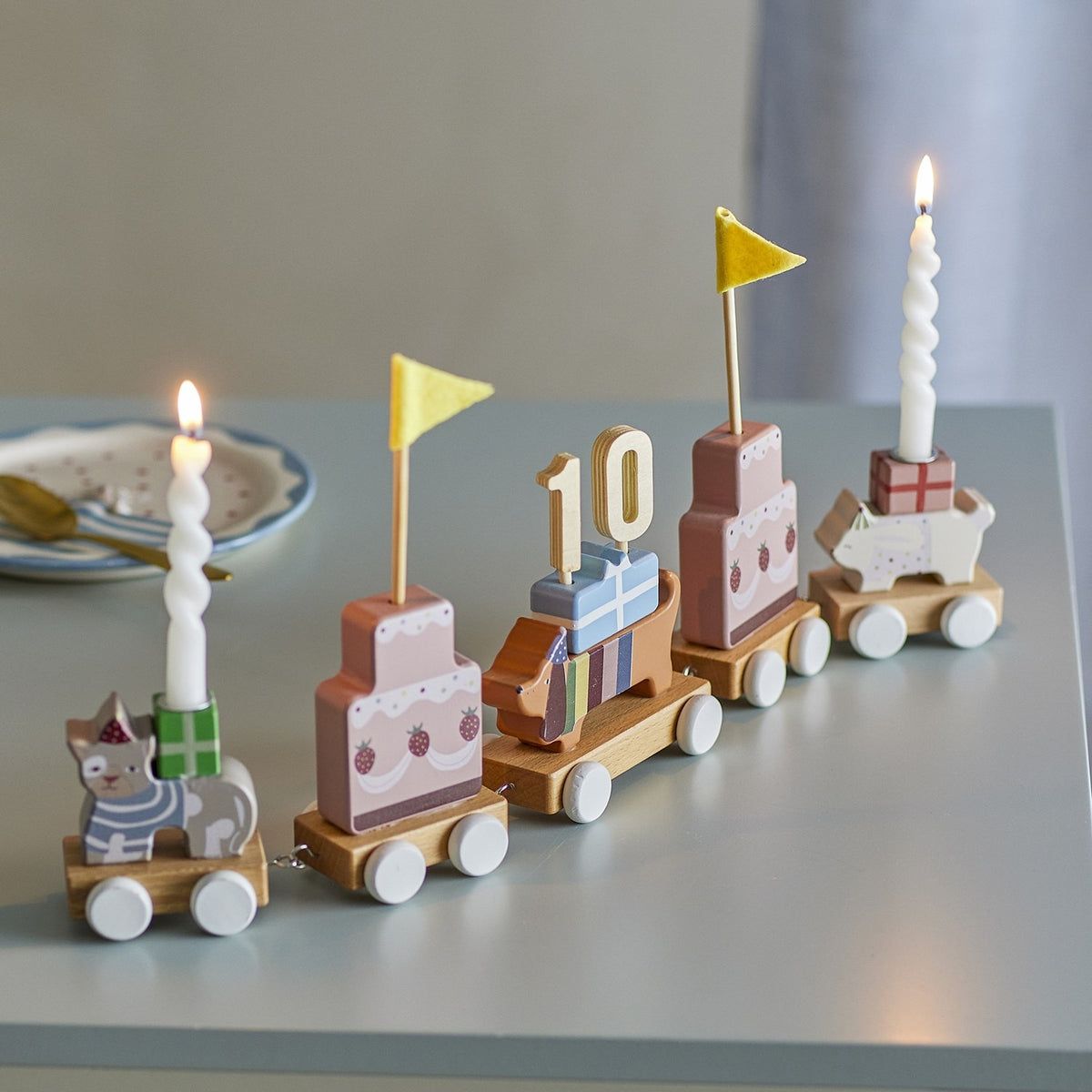 Wooden Party Birthday Decoration train with five multicoloured wagons—cat, tiered cakes, sausage dog with "10" topper—shown with lit candles on a grey table.
