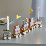 Wooden Party Birthday Decoration train with five multicoloured wagons—cat, tiered cakes, sausage dog with "10" topper—shown with lit candles on a grey table.