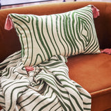 Woodsby Cotton Cushion in cream with a green woodgrain-style swirl pattern and pink corner tassels, shown on a tan velvet sofa with a matching striped throw.