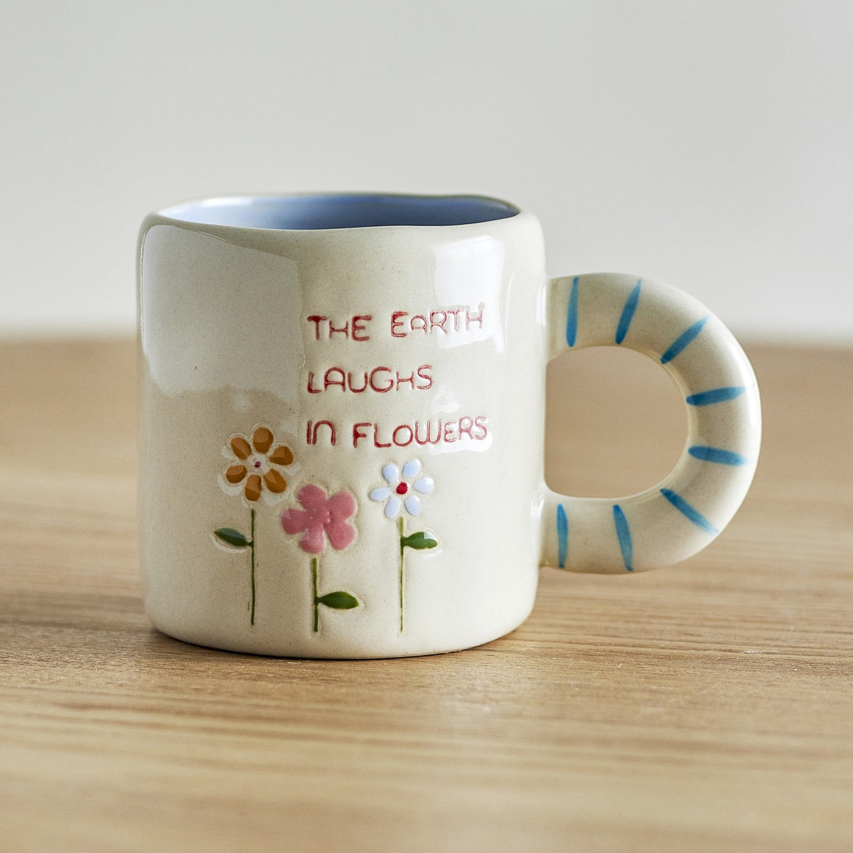 Cream stoneware cup with blue-striped ring handle, pale blue interior glaze, and hand-painted flowers beside "The earth laughs in flowers" text, on a wooden surface.
