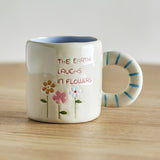 Cream stoneware cup with blue-striped ring handle, pale blue interior glaze, and hand-painted flowers beside "The earth laughs in flowers" text, on a wooden surface.