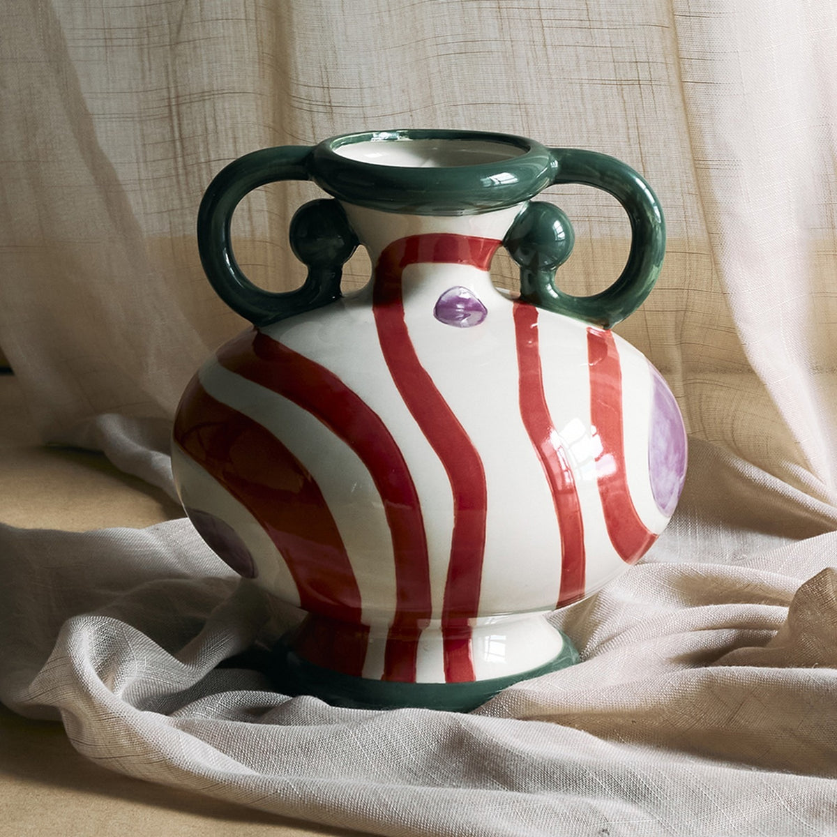 Betiva Striped Stoneware Vase 25.5cm with rounded white body, red wave stripes, purple spots, green loop handles and footed base, shown against draped fabric.
