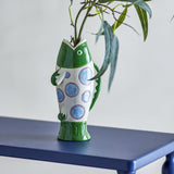 Irving Green Fish Stoneware Vase, 22cm, shaped as an upright fish with glossy green head, white body with blue spotted pattern, and flared tail base, holding leafy stems on a blue table.