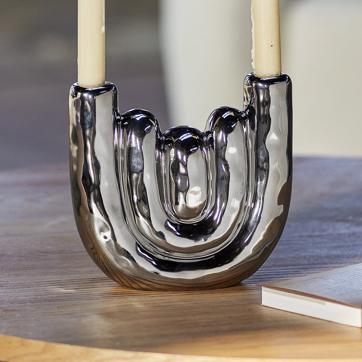 Shine Stoneware Candle Holder in silver, a U-shaped ceramic design with rippled bands and mirror-shine finish, holding two taper candles on a wooden table.