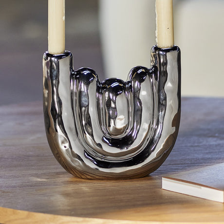 Shine Stoneware Candle Holder in silver, a U-shaped ceramic design with rippled bands and mirror-shine finish, holding two taper candles on a wooden table.