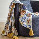 Figuria Cotton Throw in multicoloured floral, leaf and bird print with mustard tassel corners, draped over a dark grey sofa.