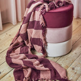 Frello Striped Cotton Throw in red, draped over a stacked velvet pouf in burgundy, pink and white, with knotted fringe trailing onto a wooden floor.