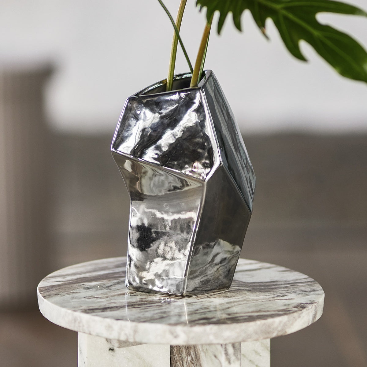 Dimona Stoneware Vase 23cm in Silver, faceted angular form with mirrored finish, shown with foliage on a marble side table.