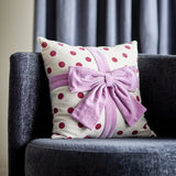 Bowie Dotted Cotton Christmas Cushion in cream with red polka dots and a mauve fabric bow, styled on a dark sofa with curtains behind.
