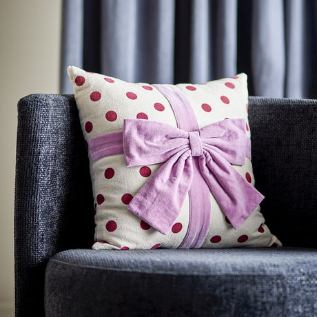 Bowie Dotted Cotton Christmas Cushion in cream with red polka dots and a mauve fabric bow, styled on a dark sofa with curtains behind.