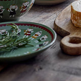 Peo Stoneware Christmas Plate in green with a scalloped red border and painted angel figures, shown with fresh rosemary on a wooden table beside cheese.