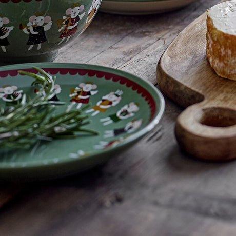 Peo Stoneware Christmas Plate in green with a scalloped red border and painted angel figures, shown with fresh rosemary on a wooden table beside cheese.