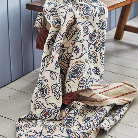 Cotton blend throw in cream with navy paisley floral print and rust tassels, draped over a wooden bench with striped reverse side visible.