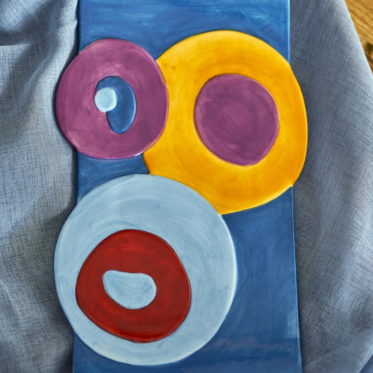 Polivia Geometric Ceramic Tray in multicoloured dolomite, hand-painted overlapping circles in purple, yellow, red and pale blue, shown on a blue surface with grey fabric backdrop.