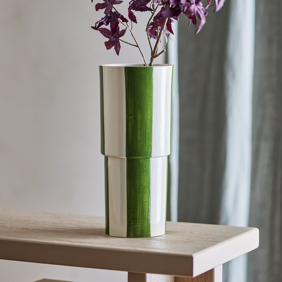 Floki Striped Stoneware Vase 31cm in green, a tall cylindrical ceramic vase with hand-painted broad stripes and a subtle stepped form, shown on a wooden table with a sprig of purple foliage.