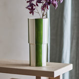 Floki Striped Stoneware Vase 31cm in green, a tall cylindrical ceramic vase with hand-painted broad stripes and a subtle stepped form, shown on a wooden table with a sprig of purple foliage.