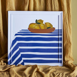 Limone Striped Wooden Framed Print 40x40cm, square wall art showing a bowl of lemons on a blue and white striped cloth, white wood frame, shown against draped gold fabric.