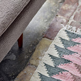 Desmond Geometric Cotton Rug in cream, pink and grey, showing a woven zigzag triangle pattern with fringed edge, placed beside a sofa leg on a stone floor.
