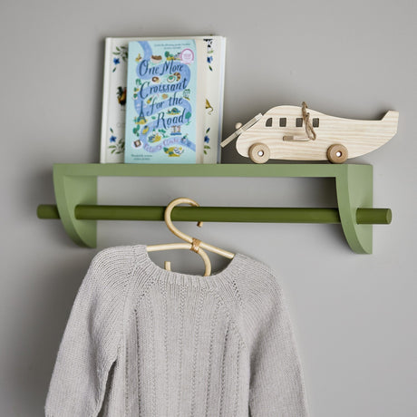Mingus wooden wall shelf in green paulownia wood with a round hanging rail, holding a book and wooden toy plane, with a grey jumper hanging below.