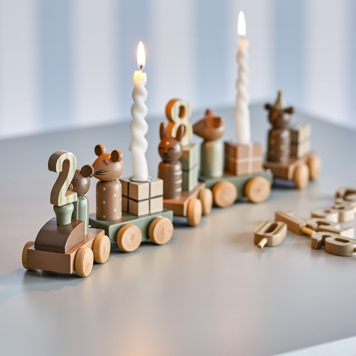 Junaid Dark Brown Birthday Decoration wooden train with number pegs, animal figures, gift block carriages and candle holders with lit spiral candles, on a light grey surface.