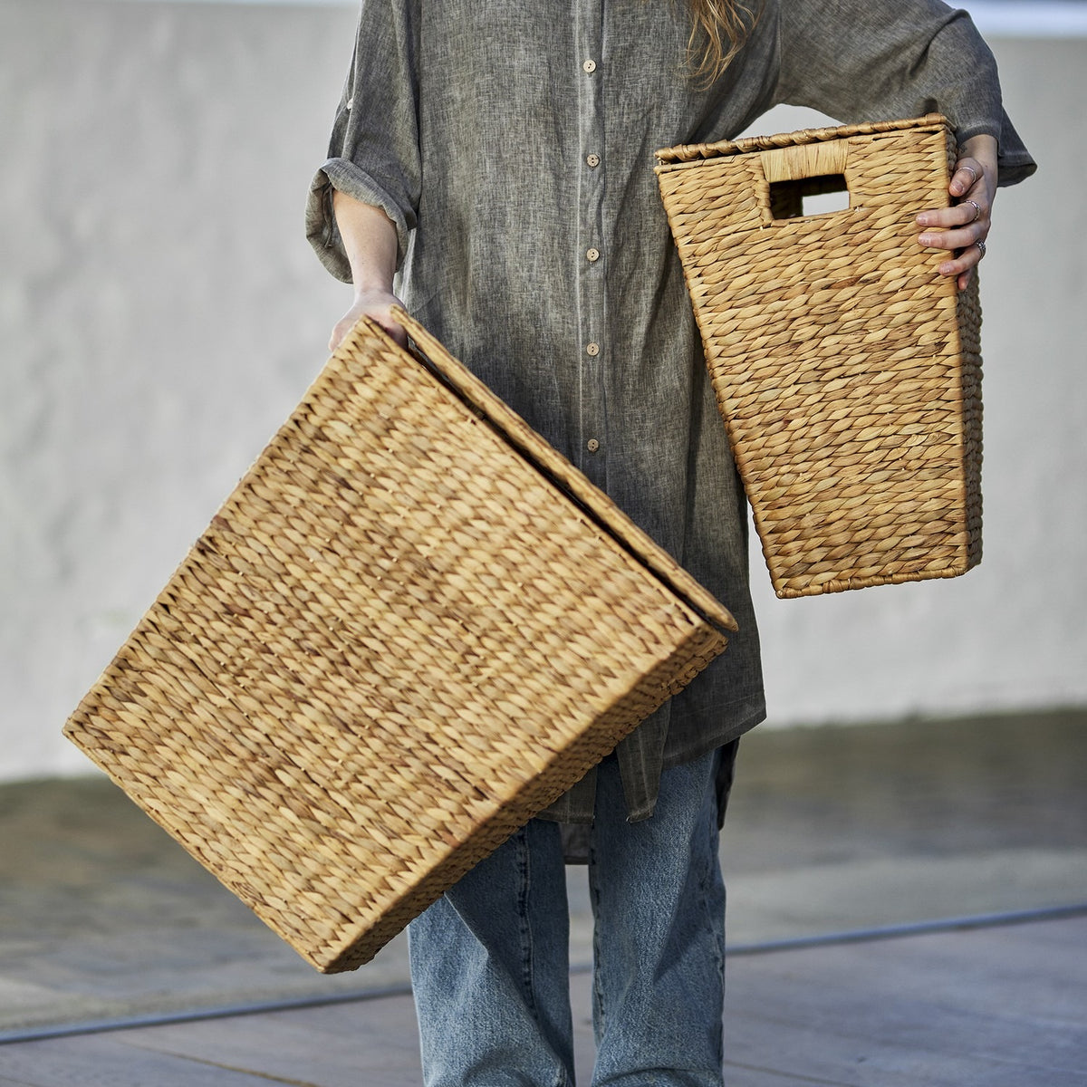 Person holding the Mios Water Hyacinth Laundry Baskets, Set of 2, showing the natural woven lids and rectangular cut-out handles on both tapered baskets.