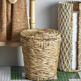 Peri Water Hyacinth Basket with Lid in natural tone, round tapered body with twisted rope weave and flat spiral lid with knotted loop, shown on patterned tile in a bathroom setting.