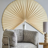 Jaime Bamboo Wall Decor 110cm in natural finish, a large pleated fan-shaped wall art mounted above a grey upholstered armchair in a living room setting.