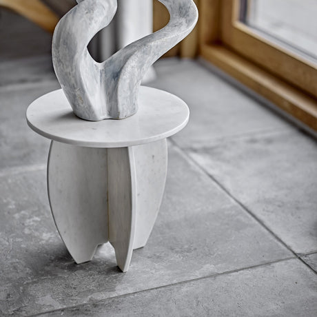 Sendai Marble Pedestal in white, a round-top side table with four curved cross-set legs, shown indoors on grey tiles with an abstract sculpture on top.