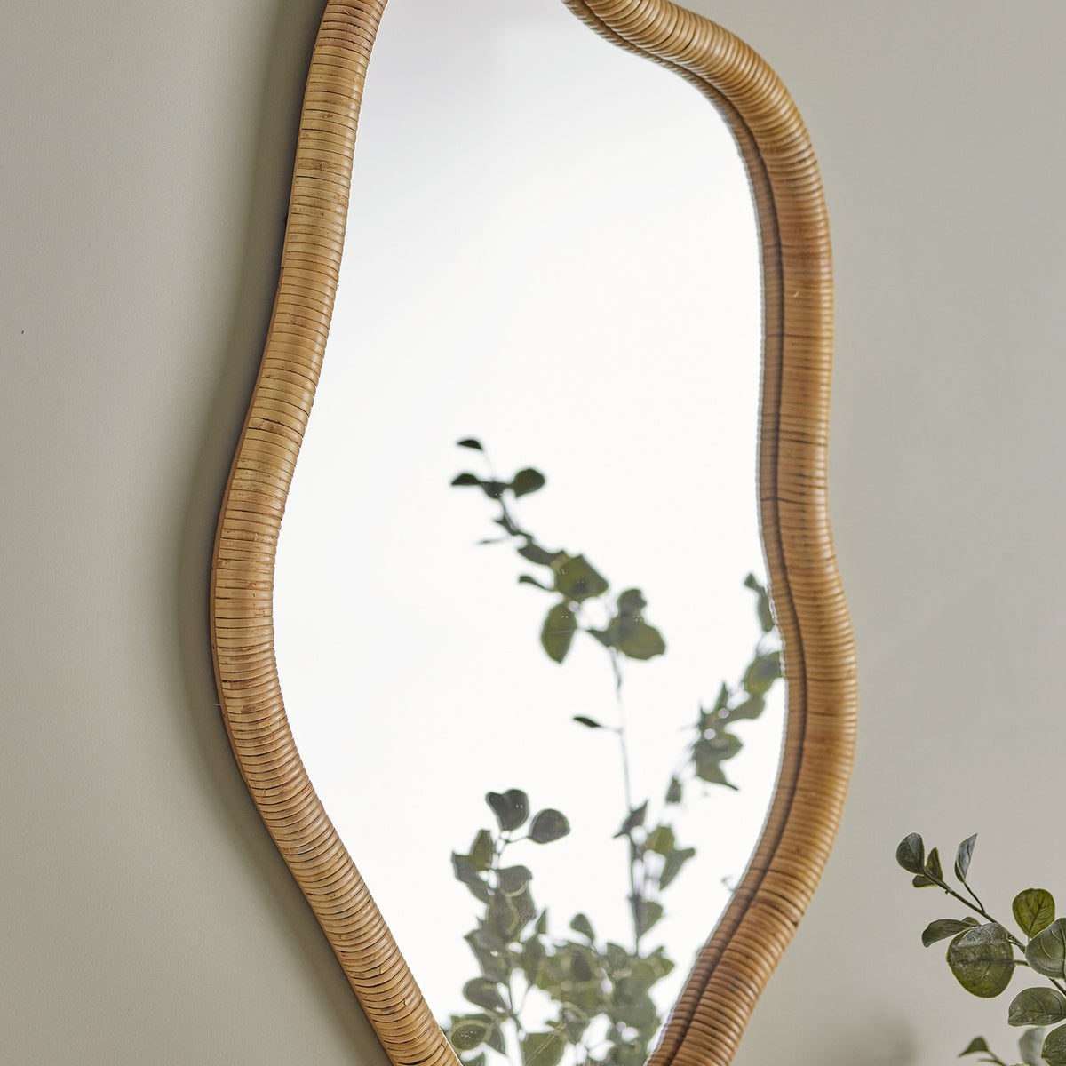 Ashlie Rattan Wall Mirror in natural rattan with a wavy, freeform frame, shown mounted on a neutral wall beside greenery.