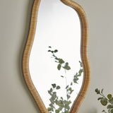 Ashlie Rattan Wall Mirror in natural rattan with a wavy, freeform frame, shown mounted on a neutral wall beside greenery.