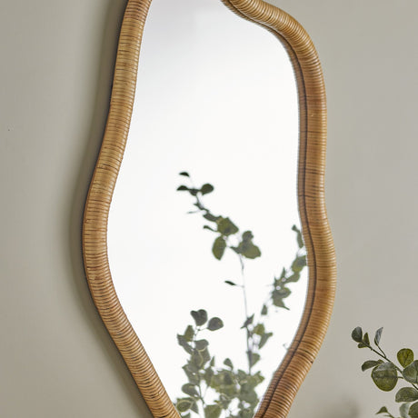 Ashlie Rattan Wall Mirror in natural rattan with a wavy, freeform frame, shown mounted on a neutral wall beside greenery.