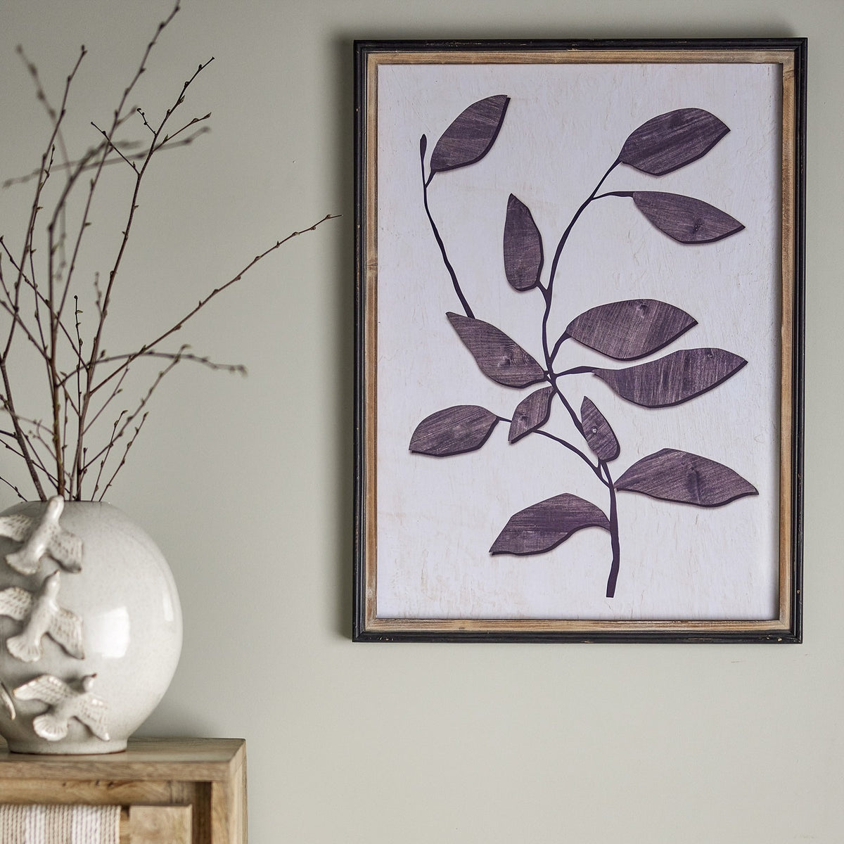 Lousa Wooden Framed Print in Black, showing a single branching leaf stem on a white ground, framed in black with an inner wood band, hung on a neutral living-room wall beside a vase of branches.