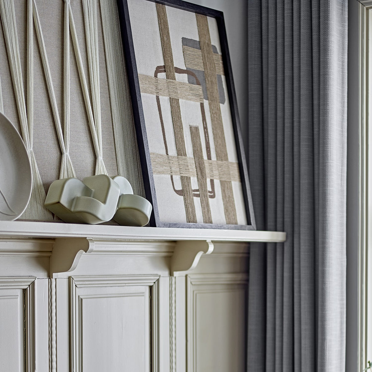 Bie Geometric Cotton Wall Decor in beige, framed textile art with woven jute bands forming a grid over rounded and square outlines, shown on a mantel shelf.