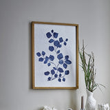 Gunnvor Wooden Wall Decor in Natural wood frame, botanical print of a navy leaf branch on textured paper, hung on a grey wall above a vase with dried grasses.