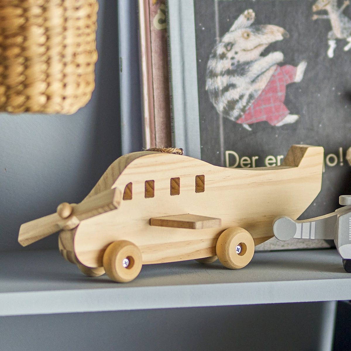 Plane Wooden Baby Mobile in natural pine with rounded nose, two-blade propeller, tail fin, side window cut-outs and wooden wheels, hemp cord loop on top, shown on a grey shelf.