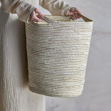 Person holding the Selena Jute Basket in White, a tall tapered storage basket with alternating white jute and seagrass stripes and two woven handles.