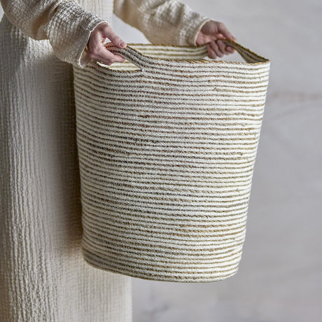 Person holding the Selena Jute Basket in White, a tall tapered storage basket with alternating white jute and seagrass stripes and two woven handles.