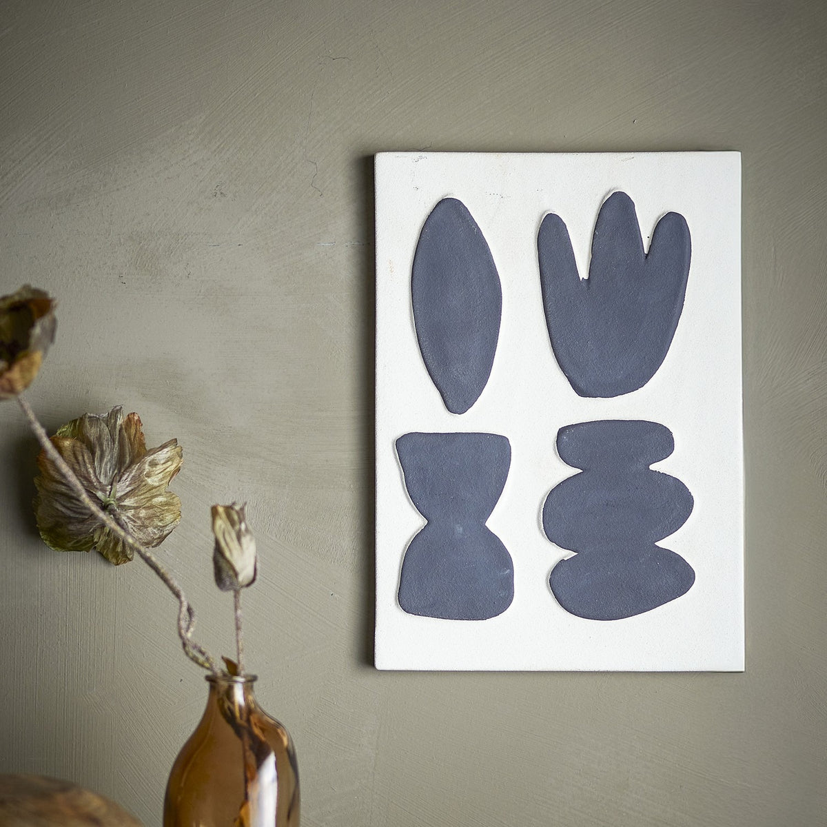 Lutton Stoneware Wall Decor in white, a rectangular panel with four raised organic shapes in grey, shown mounted on a sage green wall beside a dried floral arrangement.
