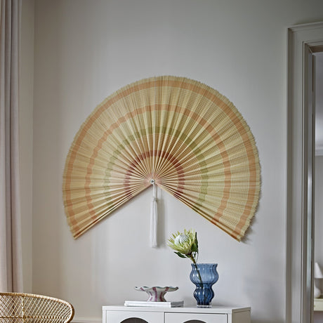 Jaime Bamboo Wall Decor in beige, a large pleated fan-shaped wall art piece with striped bands and fringed edge, hung above a console table in a living room setting.