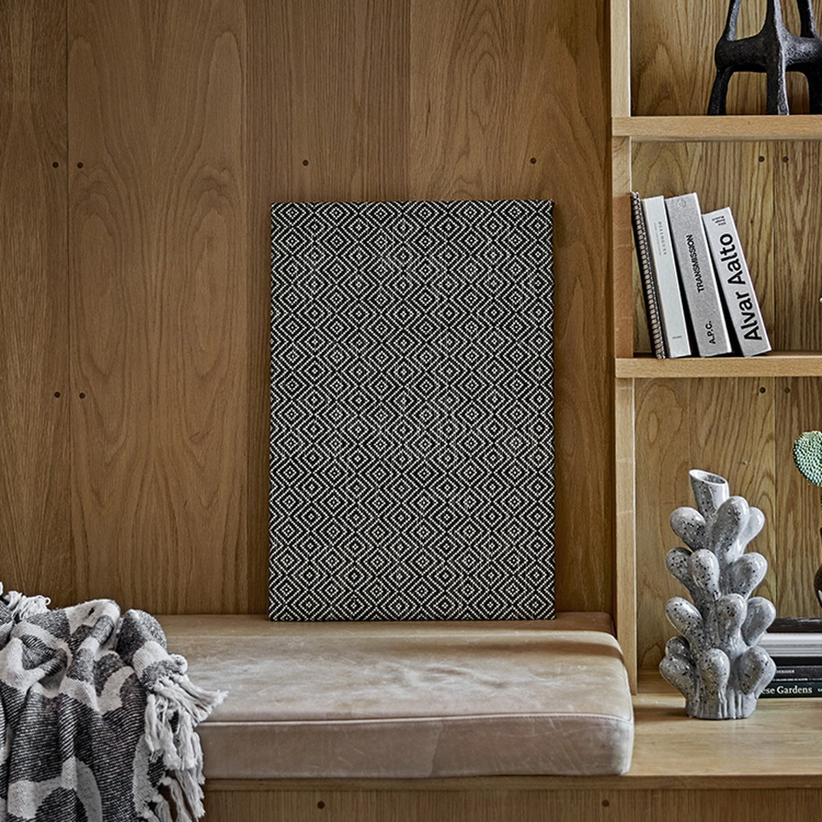 Rivalta Pin Board in black with a woven diamond pattern, leaning against a wood-panelled wall near a bookshelf and upholstered bench.