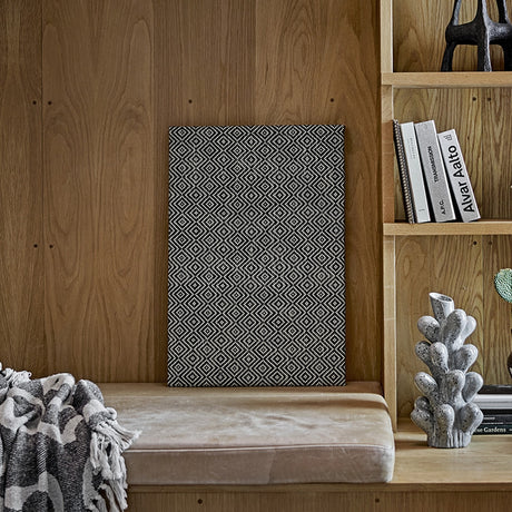 Rivalta Pin Board in black with a woven diamond pattern, leaning against a wood-panelled wall near a bookshelf and upholstered bench.
