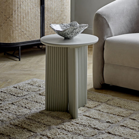 Edith Round Wooden Side Table in grey with a fluted, cross-panel base and round top, shown beside a sofa with a decorative bowl on top.