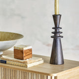 Armel Wooden Candle Holder in black, 26cm mango wood with stacked ring details, holding a taper candle on a wooden sideboard beside books and a bowl.