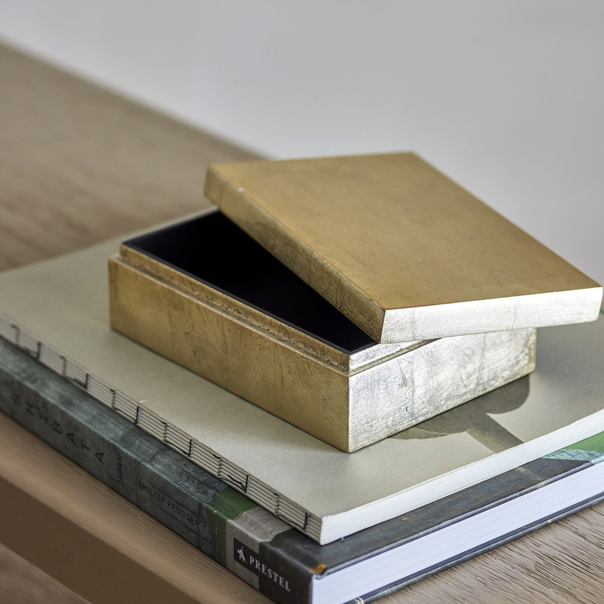 Nenon rectangular storage box with lid in gold MDF with a high-gloss finish, shown open with black interior, resting atop stacked books.