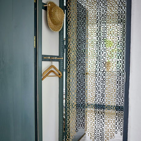 Loopi Bamboo Curtain in beige, linked ring strands hanging in a doorway beside a straw hat and wooden coat hangers in a rustic interior.