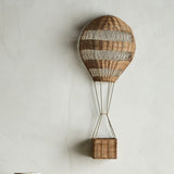 Huggi Rattan Wall Basket in natural rattan, shaped like a hot air balloon with woven striped bands and a hanging basket on a slim iron frame, mounted on a light wall.
