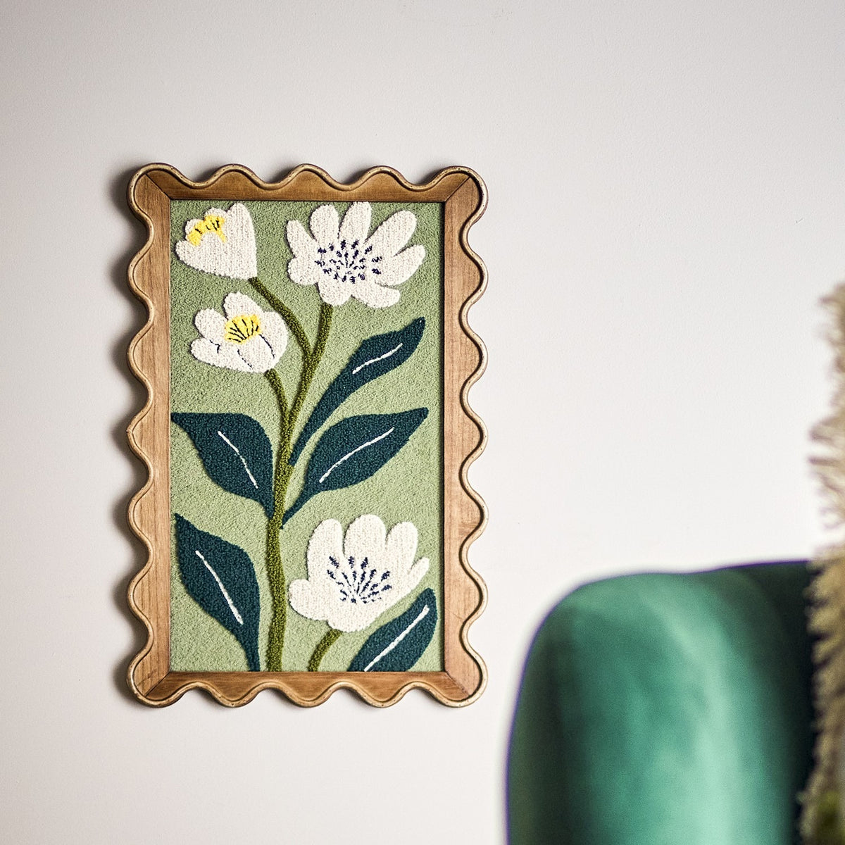 Talan Wavy Framed Print in Multi, a tufted floral wall art with white flowers and dark leaves on green, set in a wood-toned scalloped frame, shown on a white wall.